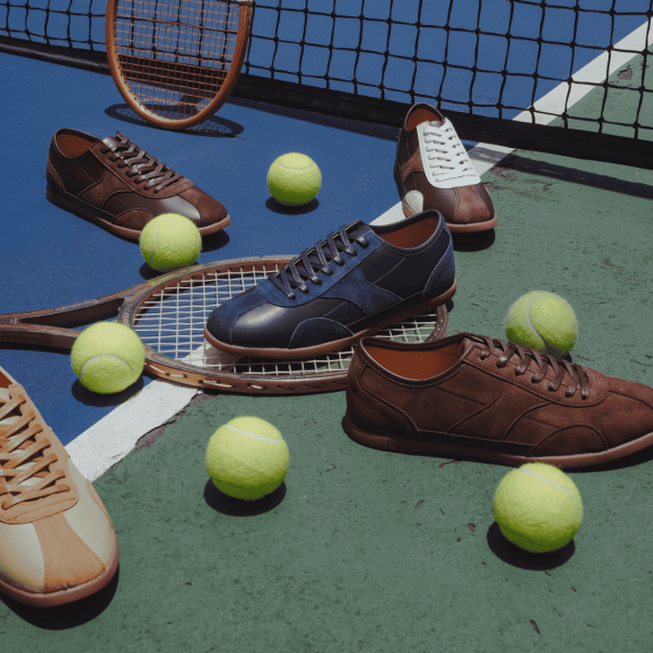 Nhiều đôi sneaker da Lucas màu nâu, xanh navy và vàng nâu đặt trên sân tennis xanh – kèm bóng tennis vàng và vợt gỗ, thể hiện sự kết hợp giữa thể thao và thời trang.